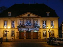 Das Bild zeigt das weihnachtlich geschmückte Rathaus von Erstein. Vor den Fenstern im ersten Stock sind kleine Tannenbäume mit Lichterketten angebracht und in der Mitte über den drei Eingangstüren im Erdgeschoss weist ein Plakat auf "Erstein un Parfum de Noel" hin.