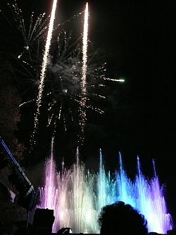 Das Bild zeigt Impressionen vom Wasserspiel "Wasserfee". Zu sehen sind lila-blaue Fontänen über denen ein weißes Feuerwerk explodiert.